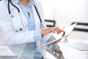 Close-up of female physician hands using digital tablet  while sitting at glass desk at hospital office. Medicine and healthcare concept