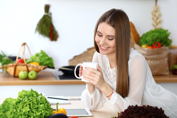 Young happy woman holding white cup and looking at the camera while sitting at wooden table in the kitchen among green vegetables. Good morning, lifestyle or cooking concept