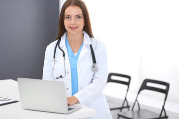 Doctor woman at work. Portrait of female physician using laptop computer while standing near reception desk at clinic or emergency hospital. Medicine and healthcare concept