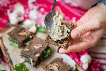 Crispy rye and flax seeds breads dehydrated and stuffed with fresh green salad leaves, mushrooms. Woman hands spread hummus paste. Vegan, vegetarian snack on Christmas red tablecloth background