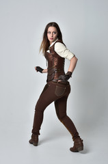 full length portrait of brunette  girl wearing brown leather steampunk outfit. standing pose with back to the camera, on grey studio background.