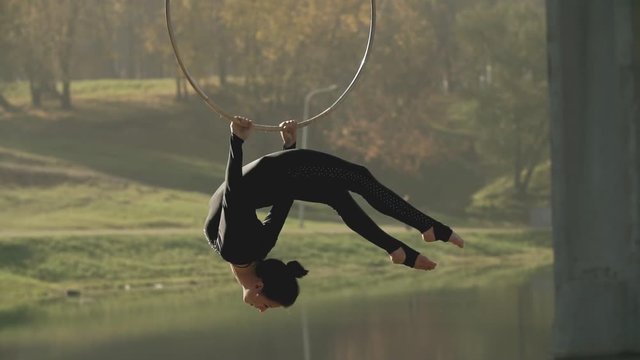 Air gymnastics woman performs acrobatics tricks on aerial hoop. Female doing some acrobatic elements on aerial hoop outdoors. Flexible brunette hanging in ring for aerial acrobatics in slow motion