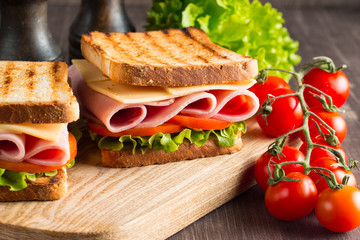 Close-up of two sandwiches with bacon, salami, prosciutto and fresh vegetables on rustic wooden cutting board. Club sandwich concept.