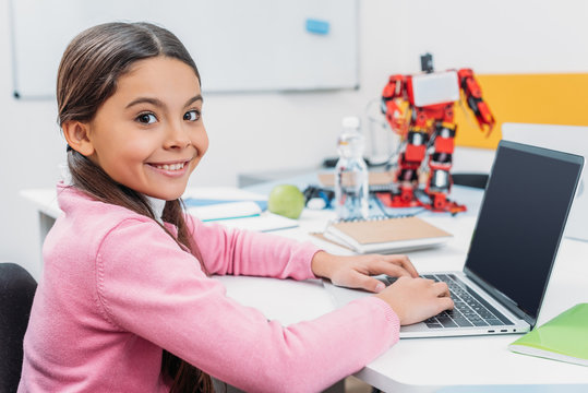 adorable schoolgirl sitting at table with robot model, looking at camera and using laptop with blank screen during STEM lesson