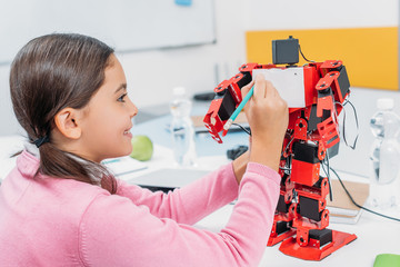 smiling schoolgirl writing with pencil on red robot chest in stem class