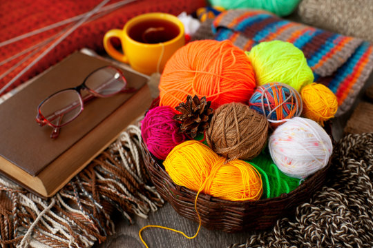 Knitting clothes in a cozy atmosphere. The yarn balls in the basket, eyeglasses, a book, a cup of tea create a coziness in the house. Knitting as a kind of hobby or needlework.