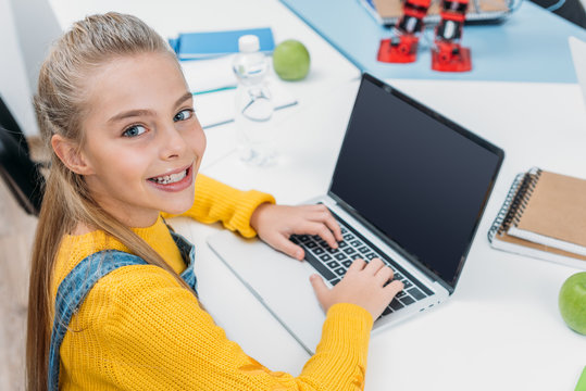 schoolgirl looking at camera and using laptop with blank screen during lesson in classroom