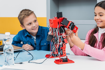 adorable schoolgirl and smiling schoolboy sitting at desk and working together on robot model at...
