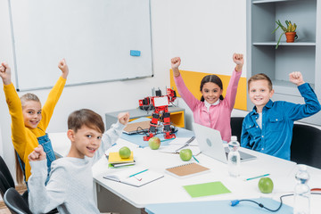 schoolchildren joying and raising hands at desk in stem education class