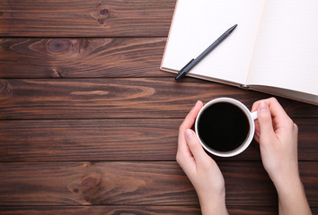Female hand holding cup of coffee on brown wooden background. Notebook and cup of coffee on brown wooden background