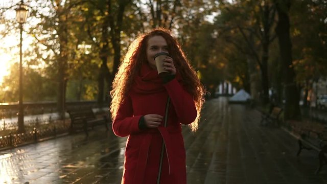 Red haired stunning woman walking on a city park. Cold weather, she wears red colored coat. Long haired beautiful, smiling girl drinks coffee from "to go" cup. Lens flares, golden sunlight. Front view