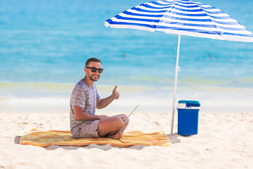 Happy young man in the beach under umbrella near the ocean working on his laptop and showing thumbs...