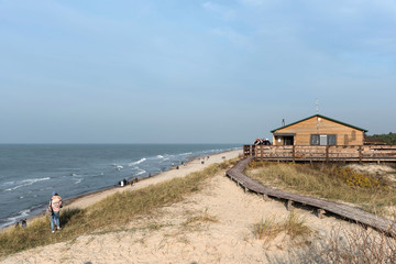 The Baltic Sea coast in the national park of Russia Curonian Spit. Tourists walk along the sea. Blue sky. To the right is a wooden walkway to the cafe. Autumn, sunny.