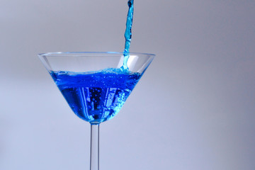 Cocktail with blue liquid in glass. Glass with blue water pouring with liquid with splashes and drops. Martini glass filling with alcohol with splashes on white background. Refreshing drink concept.