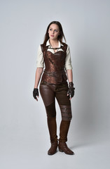 full length portrait of brunette  girl wearing brown leather steampunk outfit. standing pose on grey studio background.