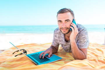 Happy smiling young man talking on mobile with laptop at summer beach