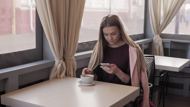 Cute girl messaging and drinking coffee in city cafe during her break. Young woman drinking coffee and relaxing alone in the morning in slow motion. Medium shot