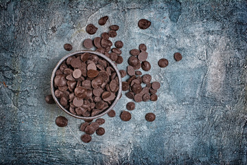 Top view of dark chocolate drops or morsels in bowl on blue concrete background