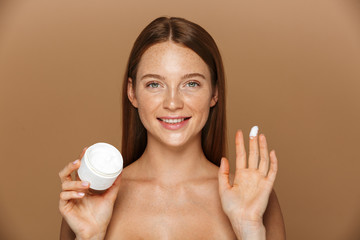 Beauty image of young shirtless woman smiling and holding jar with face cream, isolated over beige...