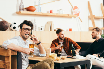 selective focus of man with beer talking on smartphone with friends behind in cafe