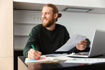 Young bearded man sitting at the kitchen writing notes in documents using laptop computer.