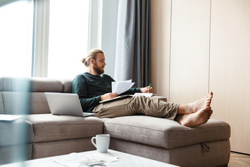 Concentrated young bearded man sitting in home work with documents.