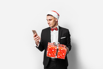 Elegant guy in a suit and santa hat, in his hands a Christmas present, looking into a smartphone,