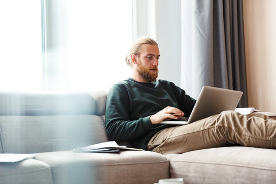 Concentrated young bearded man sitting in home using laptop computer.