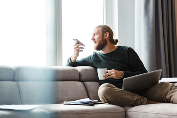 Young bearded man sitting in home using laptop computer and mobile phone.