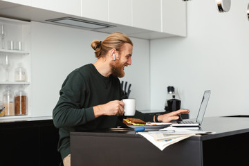 Handsome young bearded man sitting at the kitchen drinking coffee using laptop computer.