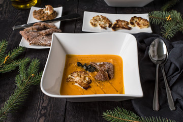 Zupa dyniowa podana z pieczonym kalafiorem, pestkami dyni i pieczywem. Ciemny drewniany stół, świąteczny wystrój.
