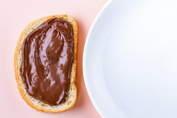 Piece of loaf with chocolate paste and a plate on a pink background. Sandwich with nut paste on a breakfast. Top view. Copy space