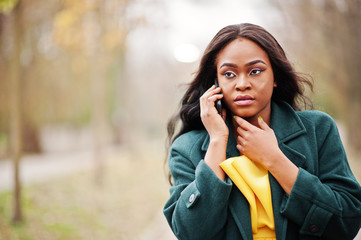 Stylish african american woman at green coat and yellow dress posed against autumn park and speaking on phone.