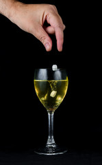 A hand dropping pills into a glass of wine, on a black background. There are two pills being dropped, one is in the wine, the other is still falling. 