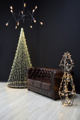 New year's Loft-style decor against a black wall, a Christmas tree from a garland. black chandelier with incandescent lamps and brown sofa. Beautiful New Year's decor with lighting in the studio