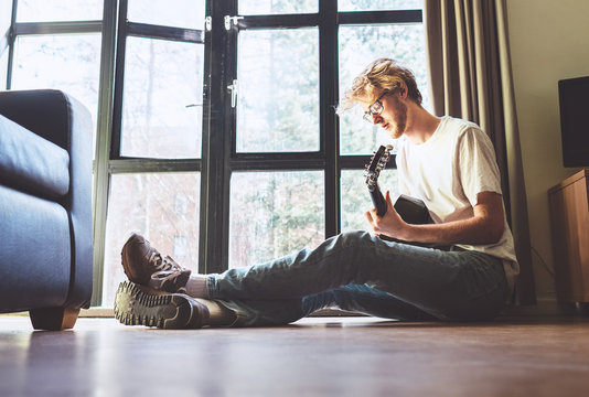 Middle shot of young man plays on guitar sitting on the floor in living room.