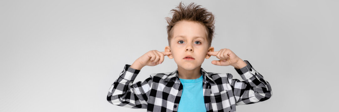 A Handsome Boy In A Plaid Shirt, Blue Shirt And Jeans Stands On A Gray Background. The Boy Folded His Arms Over His Chest. The Boy Covered His Ears With His Fingers