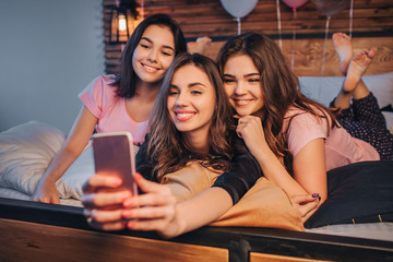 Three young models taking selfie. They pose and smile. Gils are in room. One of them hold phone and take picture on it. Young women are cheerful.
