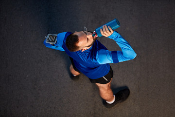 Top view of young bearded runner standing on the street in sportswear and drinking water. Around arm smart phone case.
