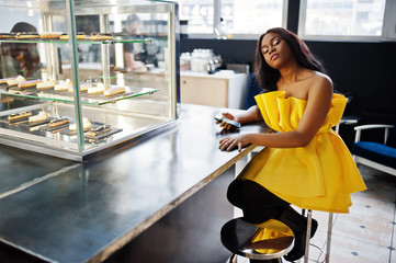Stylish african american woman at yellow dreess posed at cafe with mobile phone at hand.