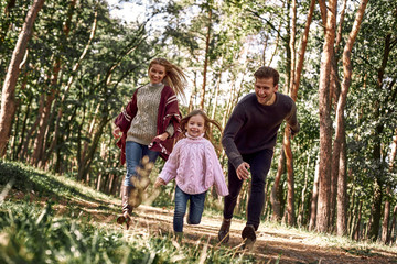 Naklejka na ściany i meble Happy parents with their daughter hold hands and run at forest path