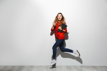 Horizontal image of beautiful woman 20s wearing red sweatshirt walking and holding gray laptop, isolated over white background