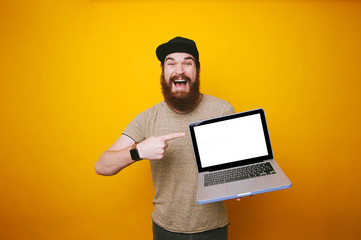 Amazed man with beard pointing at blank screen on laptop over yellow background