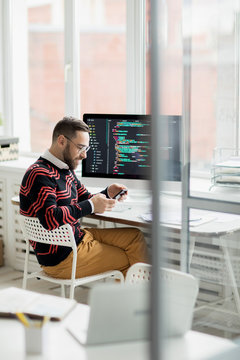 Smiling content handsome professional programmer in fashionable sweater sitting at table with modern computer and using smartphone while working in office of programming company