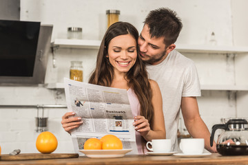 smiling girlfriend reading newspaper and boyfriend hugging her from back in morning at kitchen