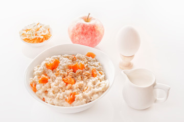 Oatmeal with pumpkin and nuts in a plate, fresh apple, vegetable salad, boiled egg and a jug with milk on a white background. Close-up. Copy space