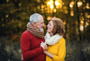 A senior couple standing in an autumn nature at sunset, looking at each other.