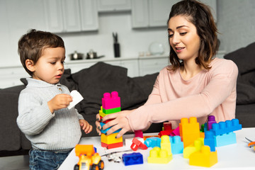 attractive happy woman and her little son playing with colorful plastic blocks at home