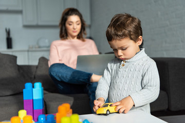  child playing with toy car while his mother using laptop behind at home