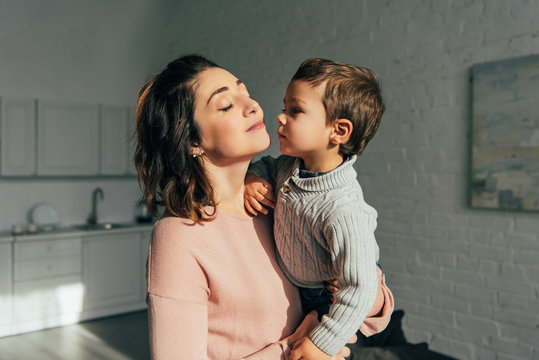 boy kissing mother while she holding him on hands at home